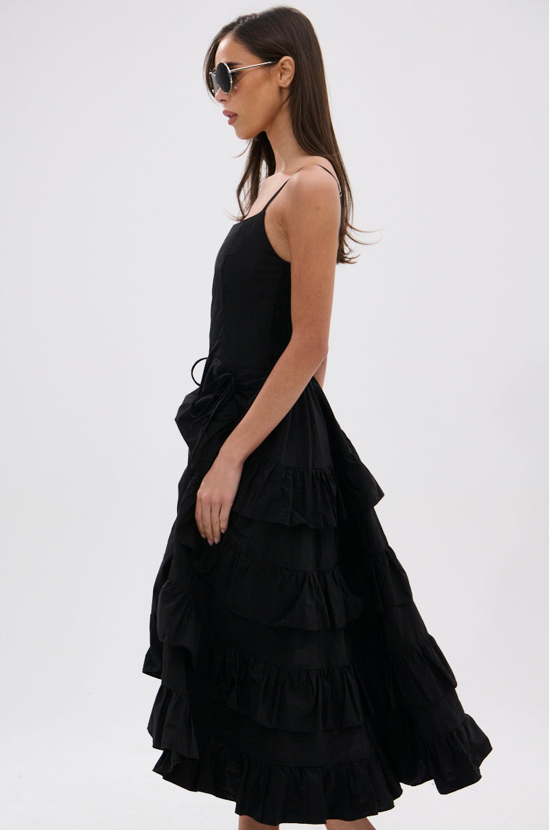 A woman in sunglasses and the ADDED DRAMA RUFFLED MIDI DRESS IN BLACK, featuring adjustable spaghetti straps, stands in profile against a plain white background.