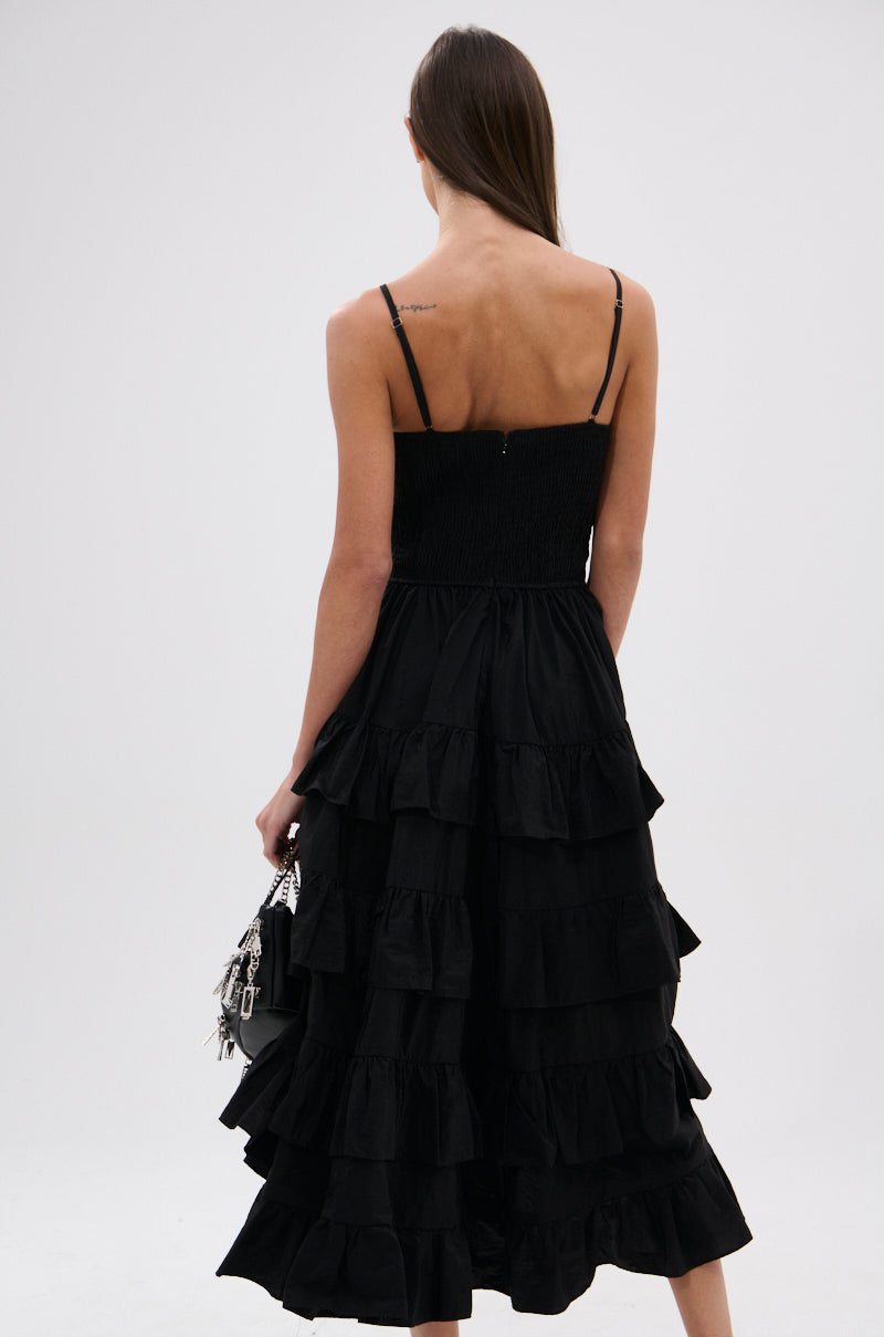 A woman with straight brown hair stands facing away, wearing the ADDED DRAMA RUFFLED MIDI DRESS IN BLACK with adjustable spaghetti straps and holding a small black handbag against a plain white background.