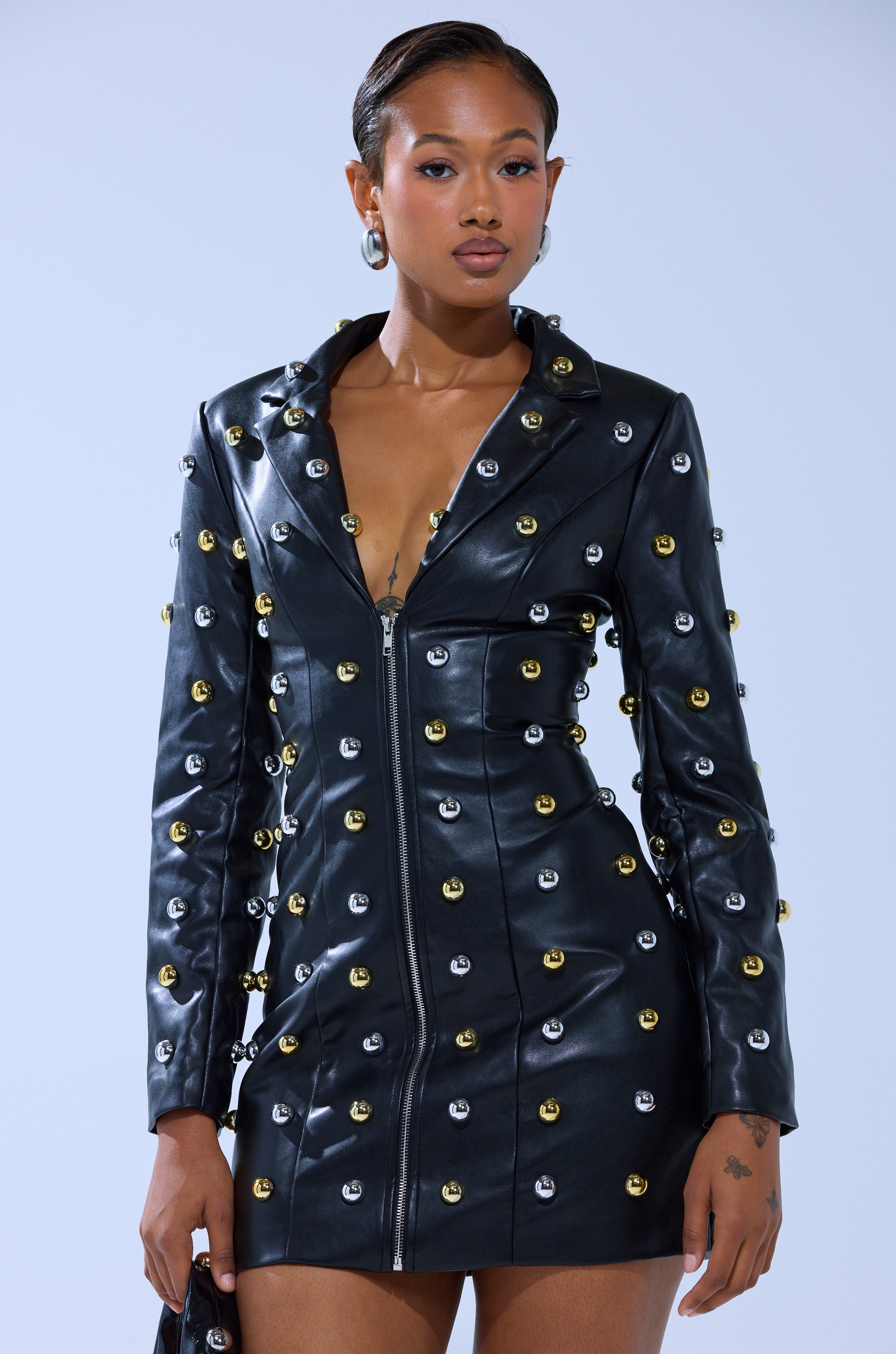 A woman stands against a plain light background, wearing the ROCK WITH IT PU BLAZER DRESS WITH HARDWARE and holding a matching studded bag.
