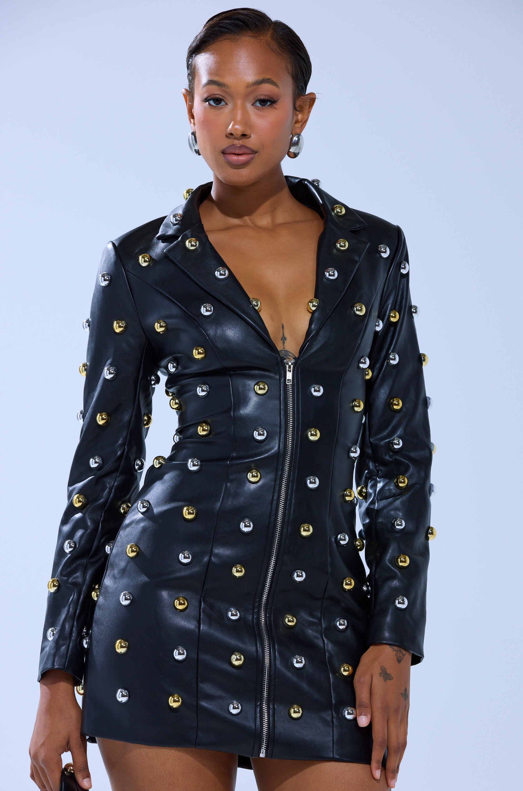 A woman stands confidently against a light background wearing the ROCK WITH IT PU BLAZER DRESS WITH HARDWARE, a fitted faux leather mini dress detailed with gold and silver metal studs. Her short, styled hair matches her calm, composed look.