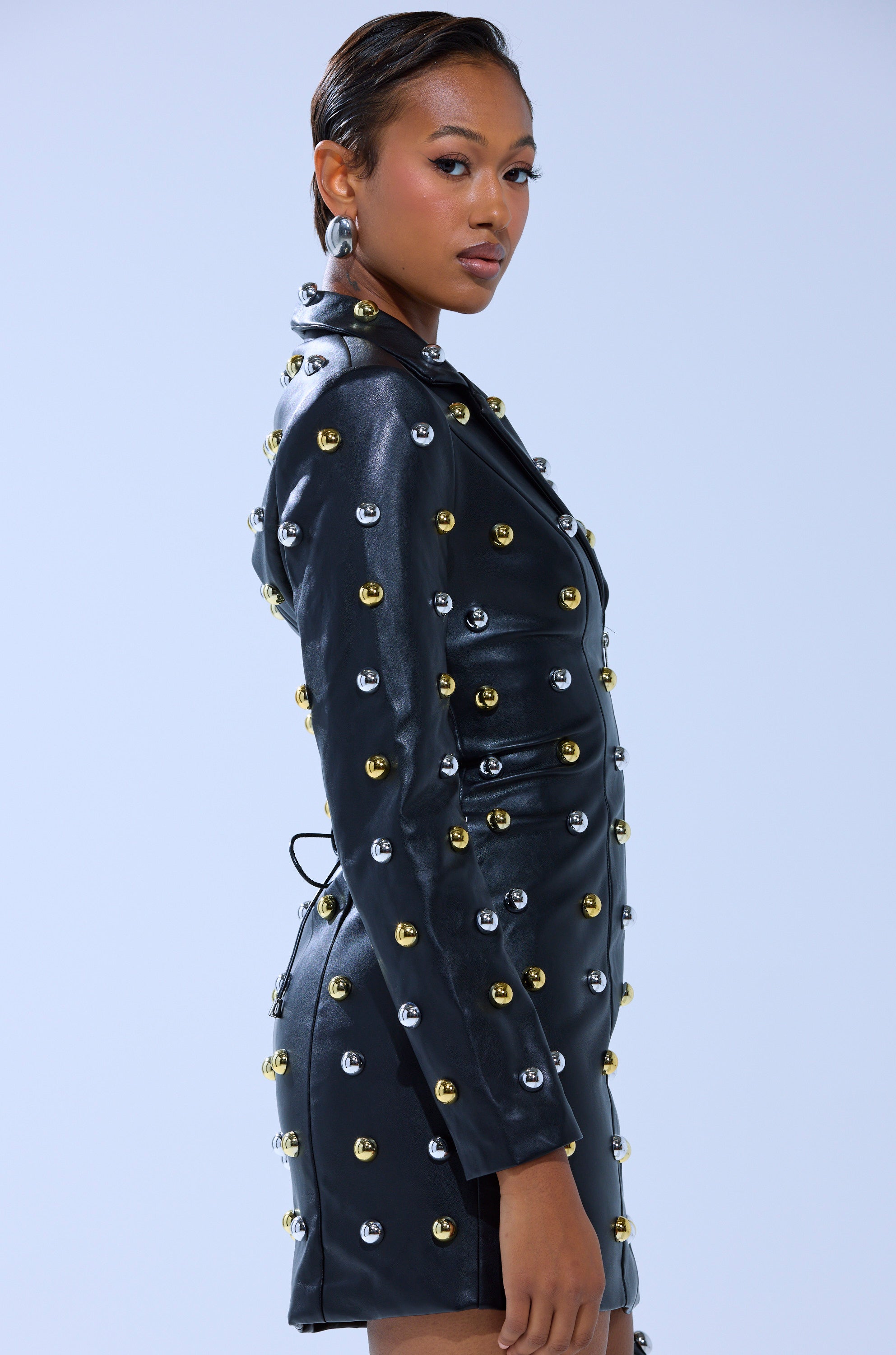 A woman stands in profile against a light blue background, wearing the ROCK WITH IT PU BLAZER DRESS WITH HARDWARE—a black fitted mini dress with silver and gold hardware. She has slicked-back hair and hoop earrings.
