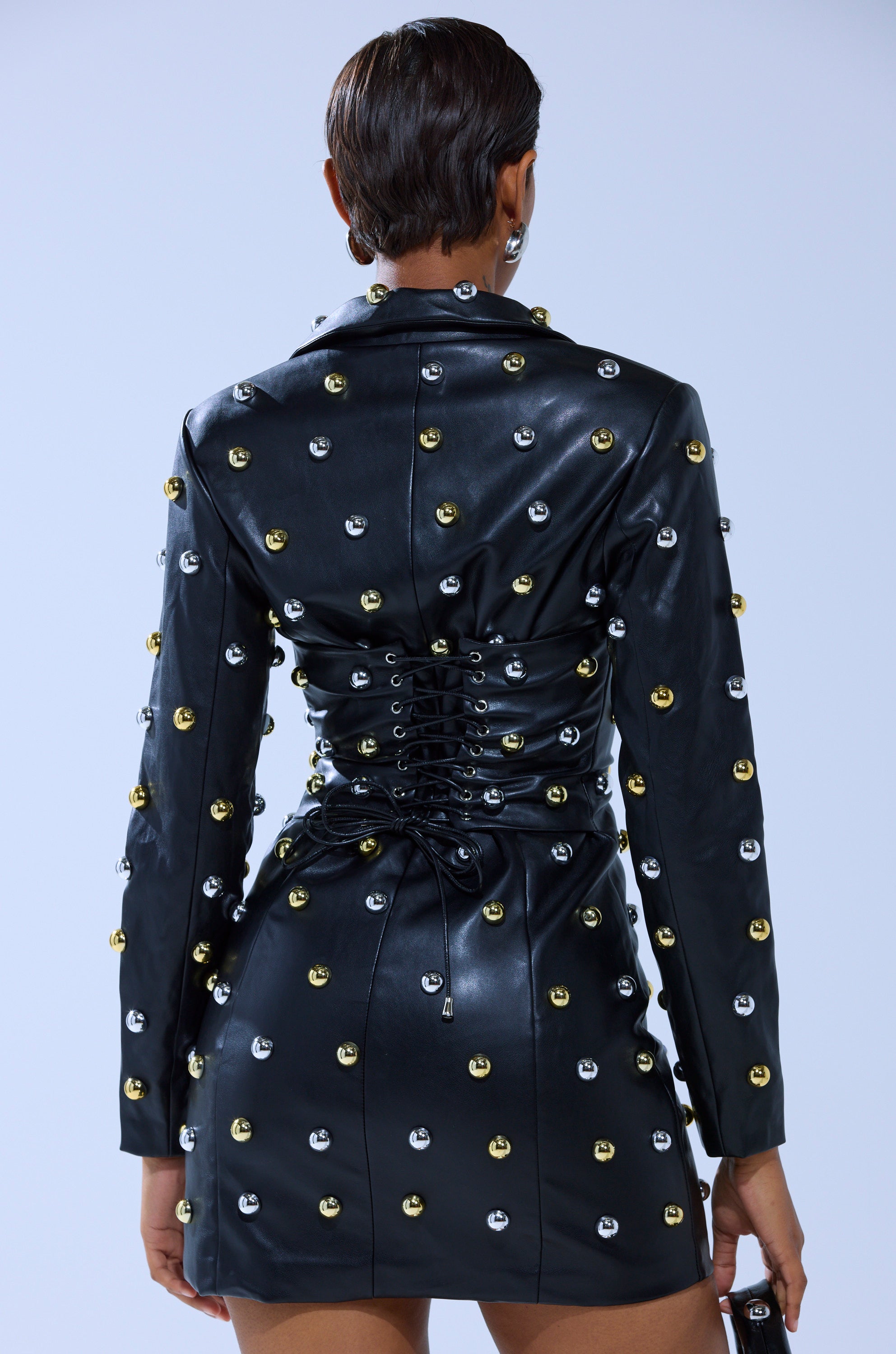 A person with short hair faces away, wearing the ROCK WITH IT PU Blazer Dress, a fitted black faux leather mini dress with metallic silver and gold studs and a lace-up corset detail on the back.