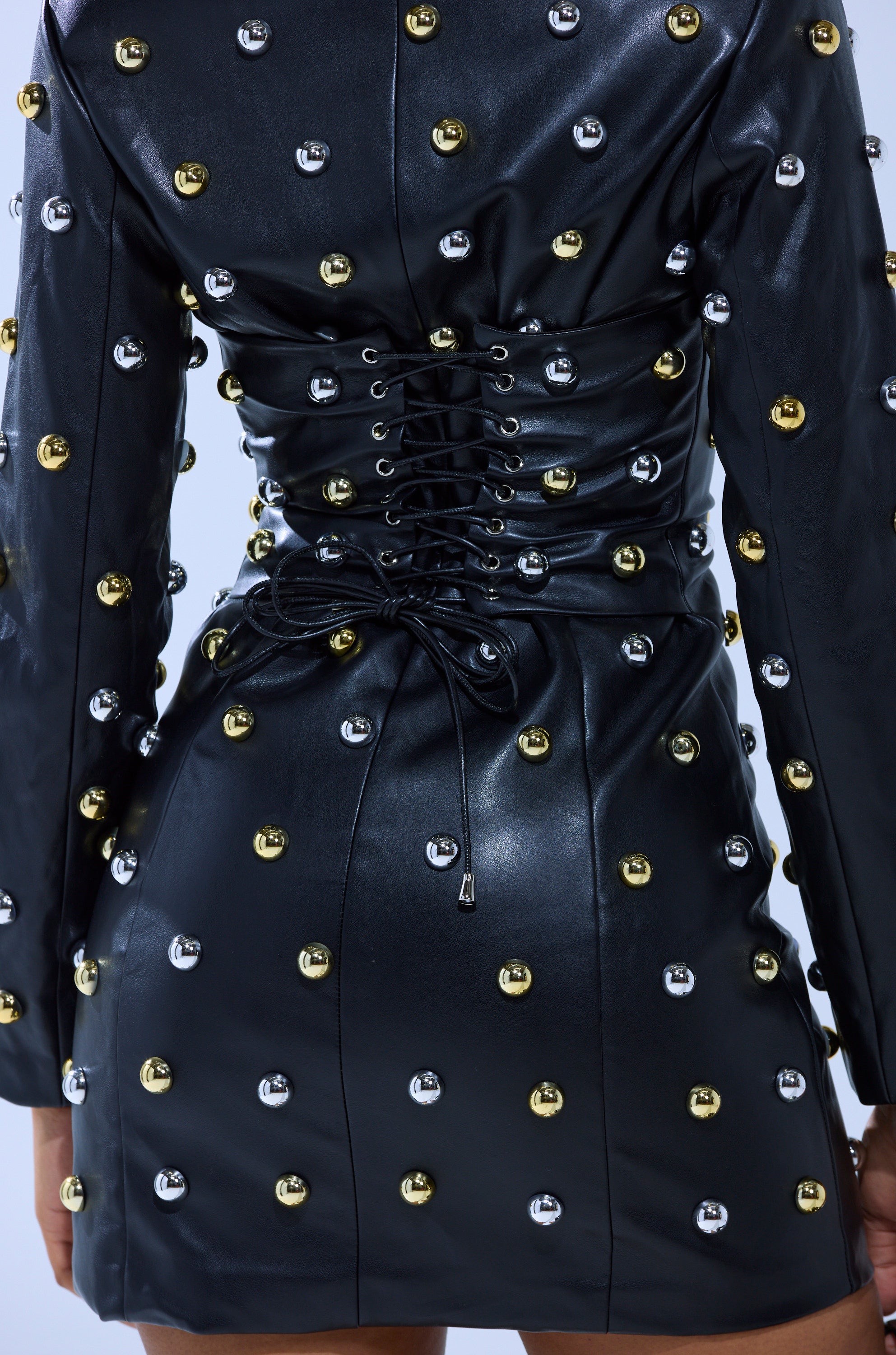 A close-up of a person wearing the ROCK WITH IT PU BLAZER DRESS WITH HARDWARE, featuring gold and silver stud embellishments, a corset-style lace-up back, long sleeves, and a short hemline.