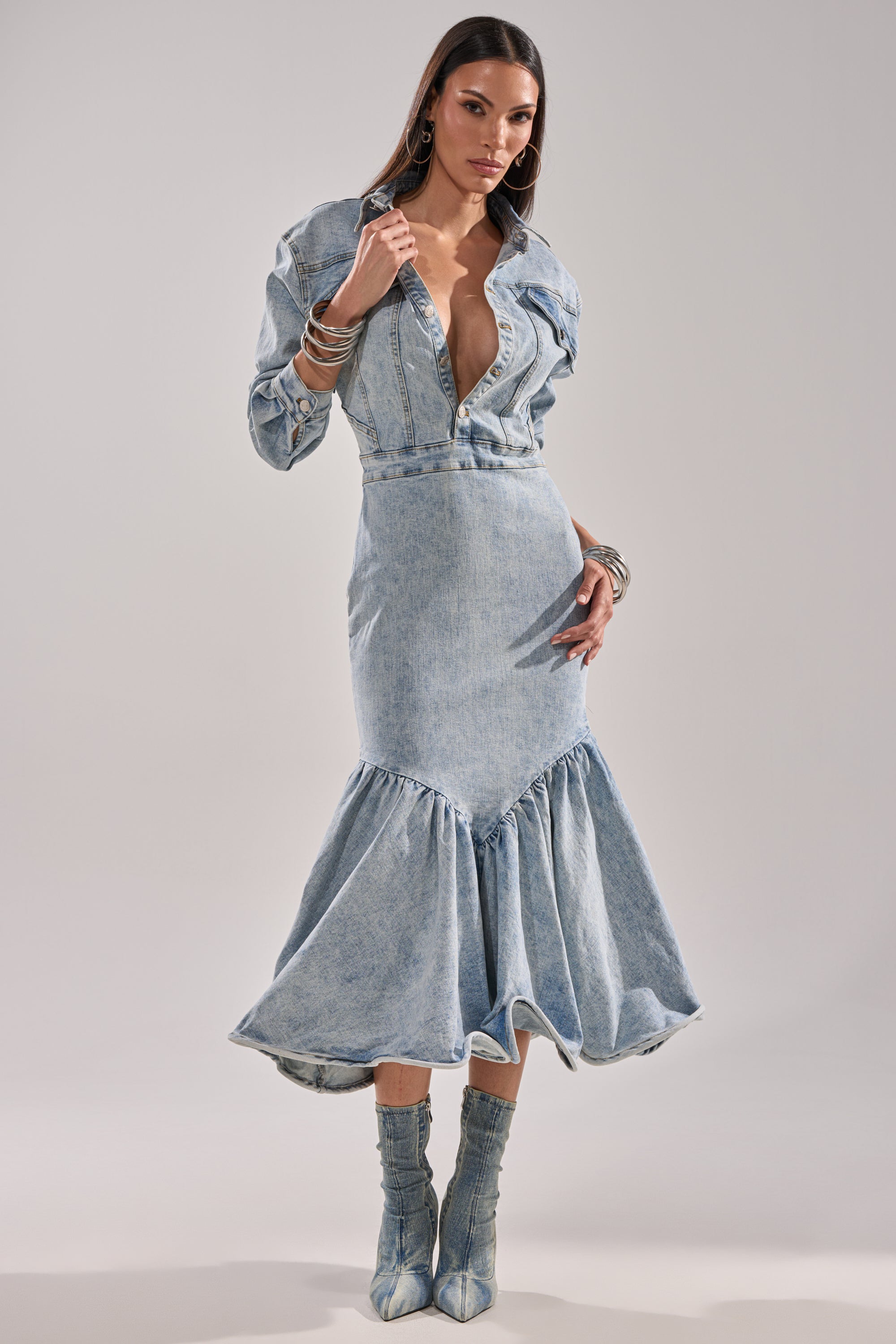 A woman models the DENIM FOR DAYS MAXI DRESS, a light-wash denim maxi with deep V-neck, long sleeves, and flared hem. She pairs it with denim boots, hoop earrings, and bracelets against a plain backdrop.