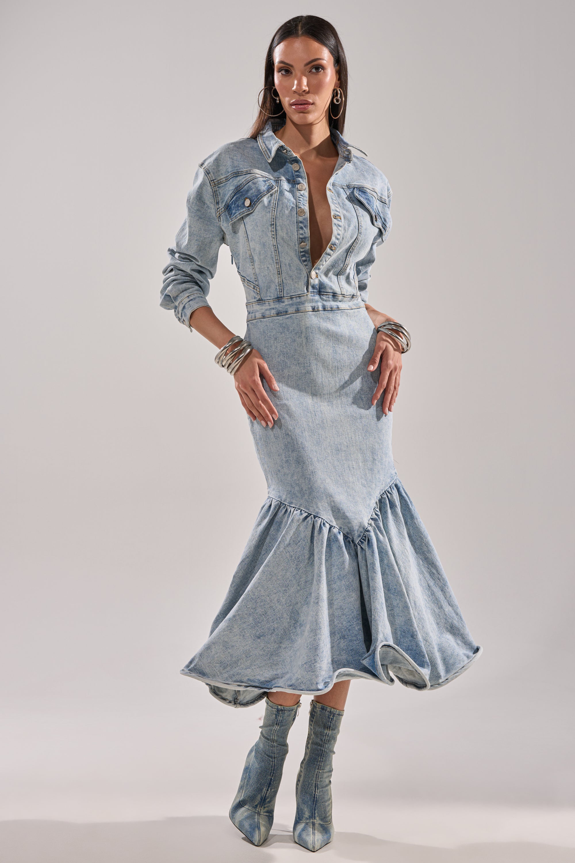A woman models the DENIM FOR DAYS MAXI DRESS, a fitted denim maxi with a flared ruffled hem, paired with light-wash denim boots and silver bangles against a plain background.