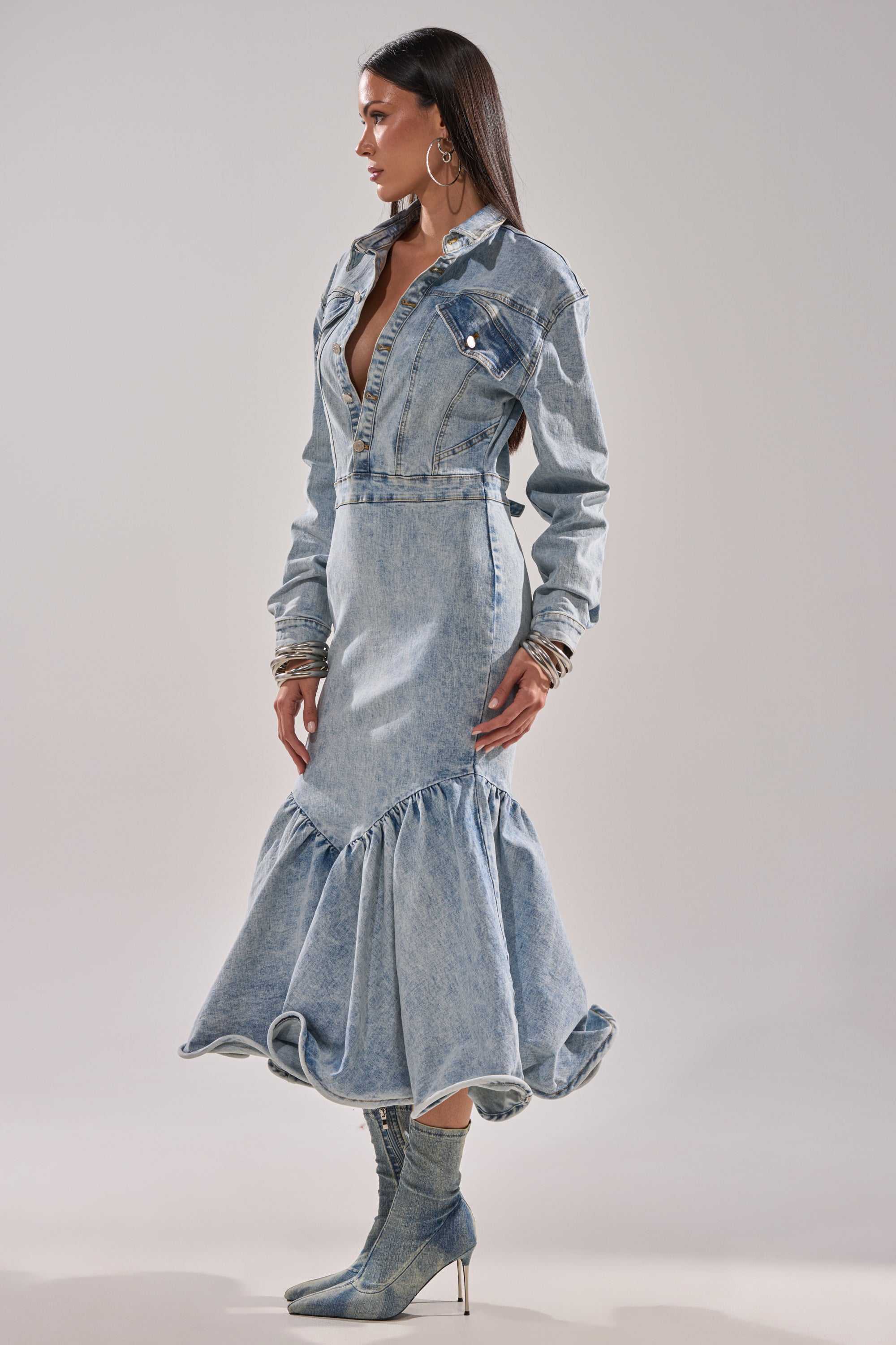 A woman models the DENIM FOR DAYS MAXI DRESS from AKIRA Label, styled with hoop earrings, bracelets, and denim heeled boots as she poses against a plain background.