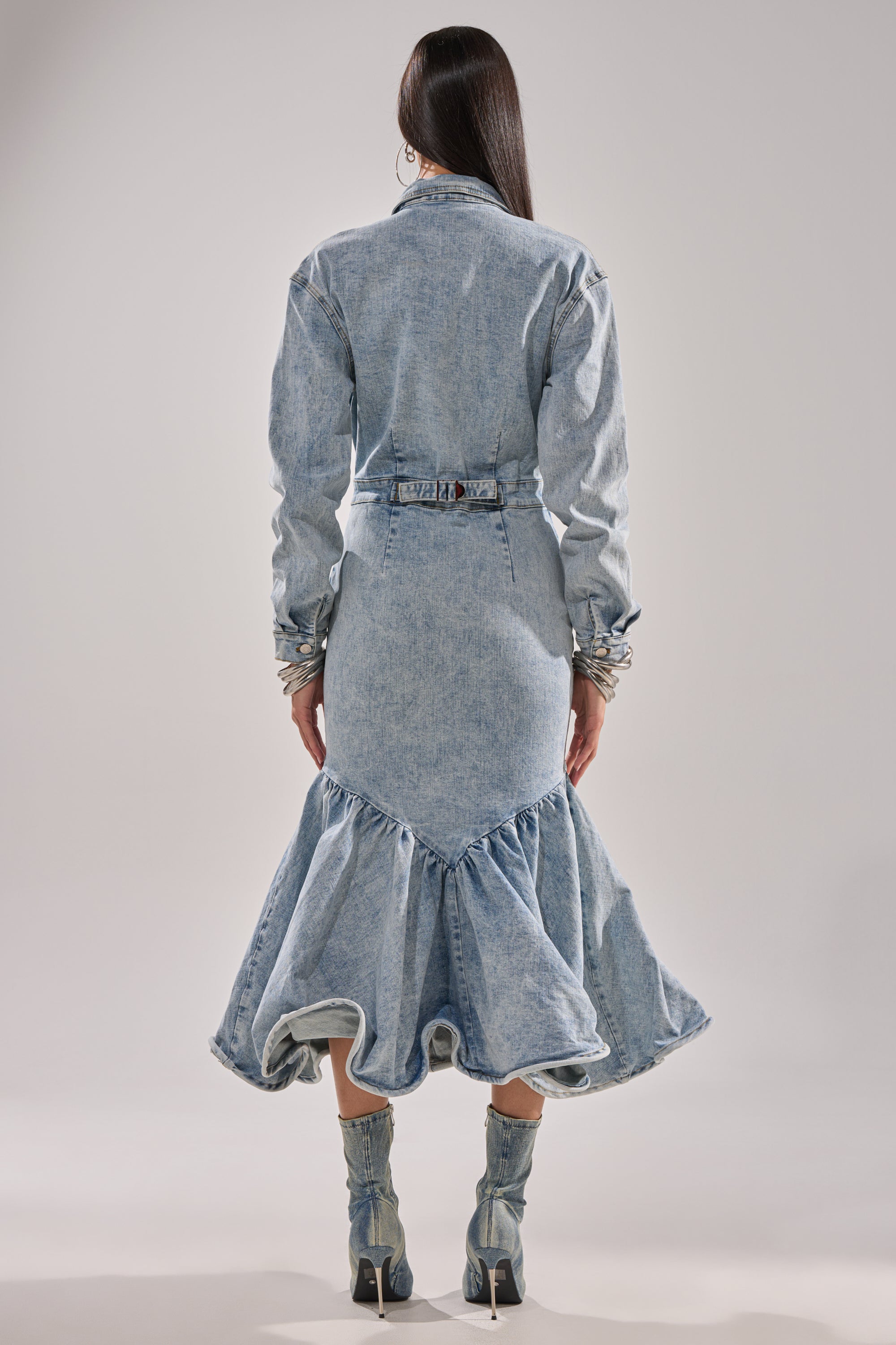 A person faces away, wearing the DENIM FOR DAYS MAXI DRESS—a light-wash denim maxi dress with a ruffled hem and belted waist—paired with matching mid-calf denim boots, set against a plain background.
