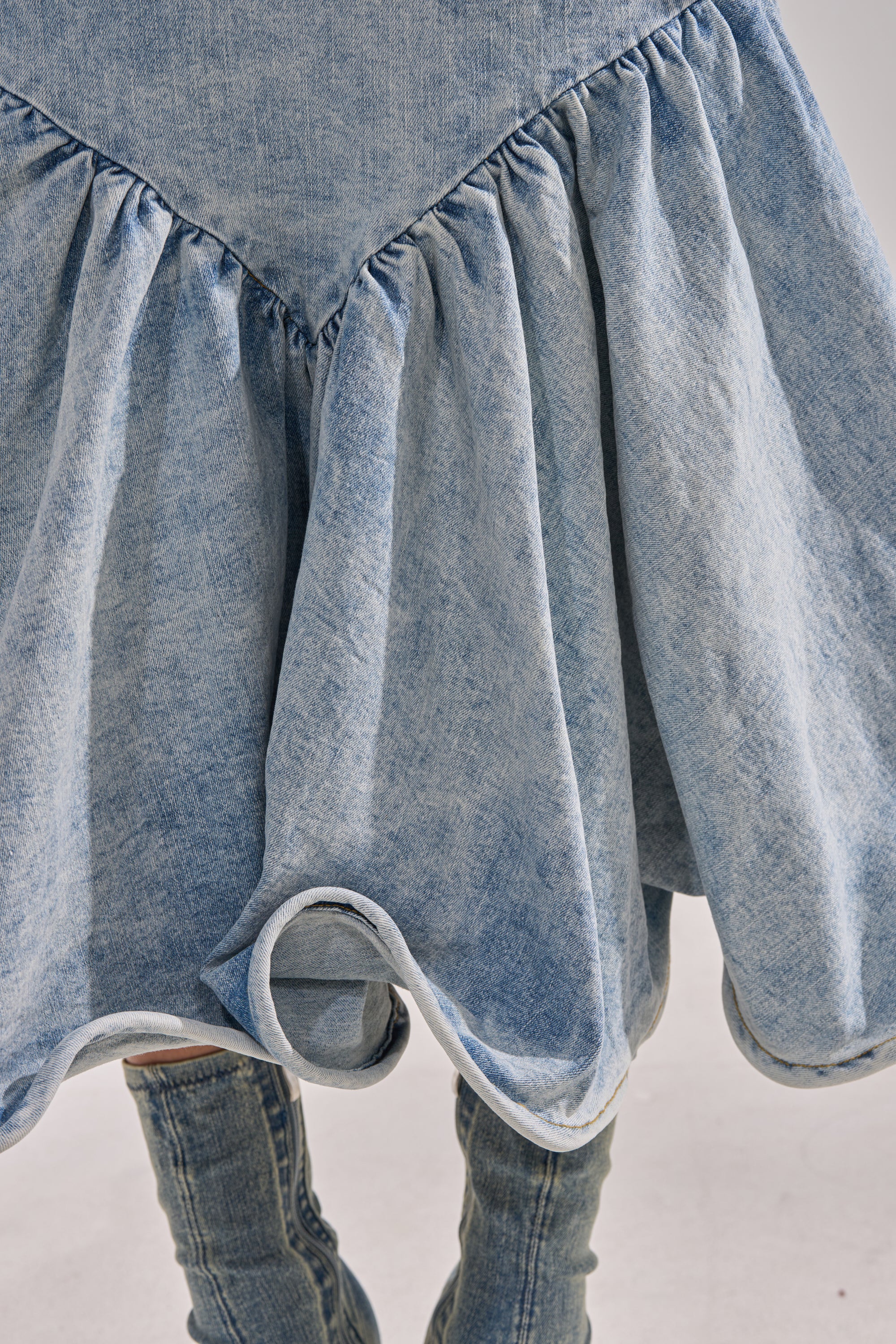 A close-up of the DENIM FOR DAYS MAXI DRESS, a curve-hugging light blue denim dress featuring ruffled hem detailing, styled with matching denim accessories against a plain light background.