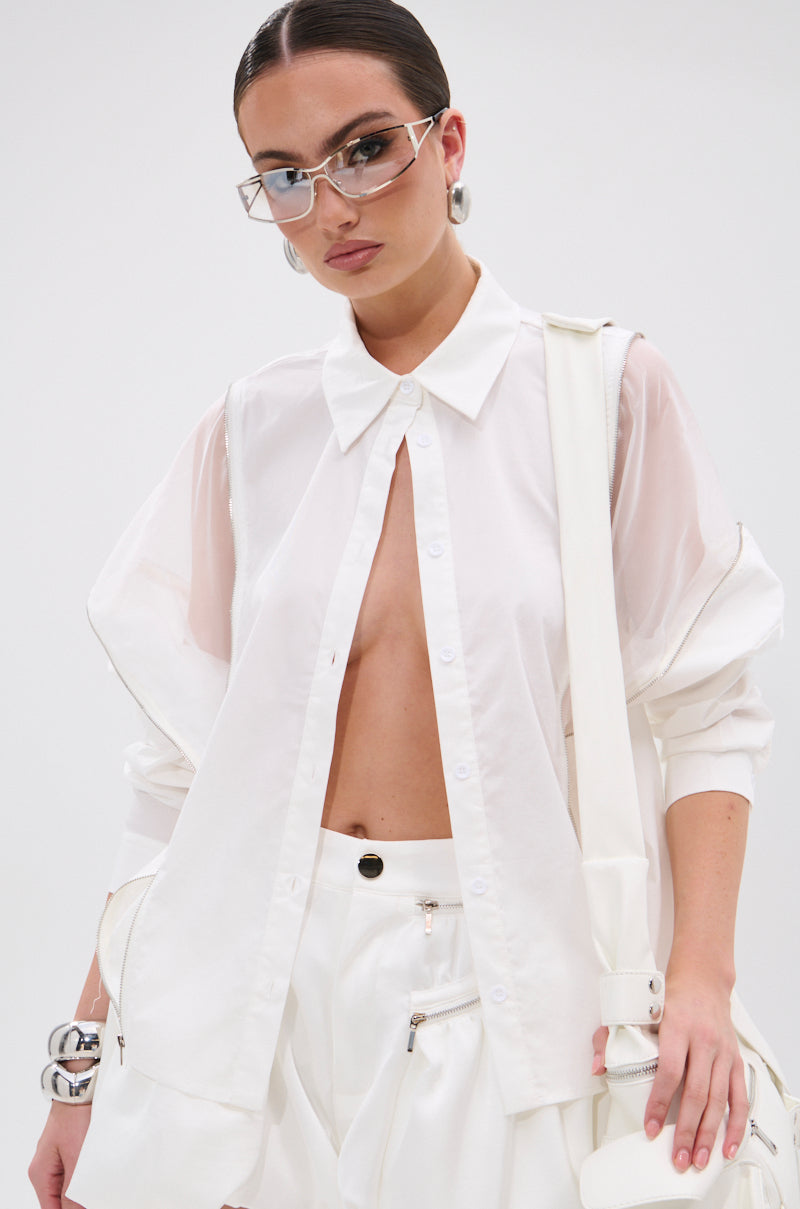 A woman with slicked-back dark hair wears the edgy UNZIP ME BLOUSE IN WHITE, left mostly unbuttoned, paired with white high-waisted pants and chunky silver jewelry against a plain light background. 1