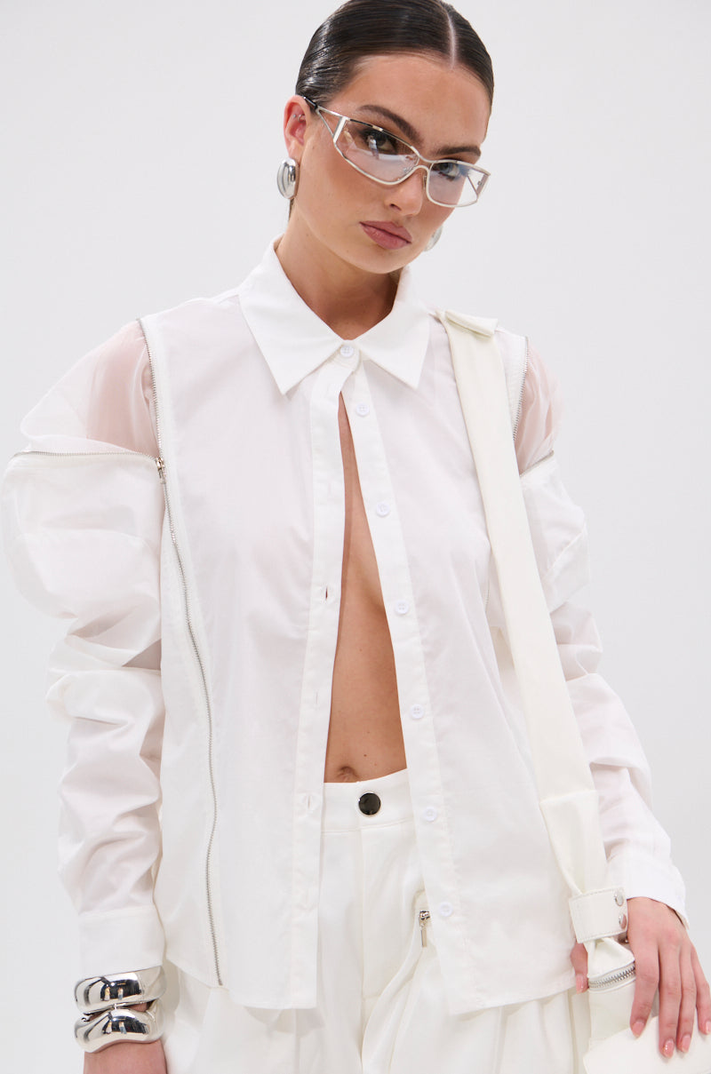 A woman models the UNZIP ME BLOUSE IN WHITE—an edgy, sheer white shirt unbuttoned to the mid-torso and fitted—paired with white pants. She accessorizes with silver bangles, clear glasses, and slicked-back hair for a bold look. 2