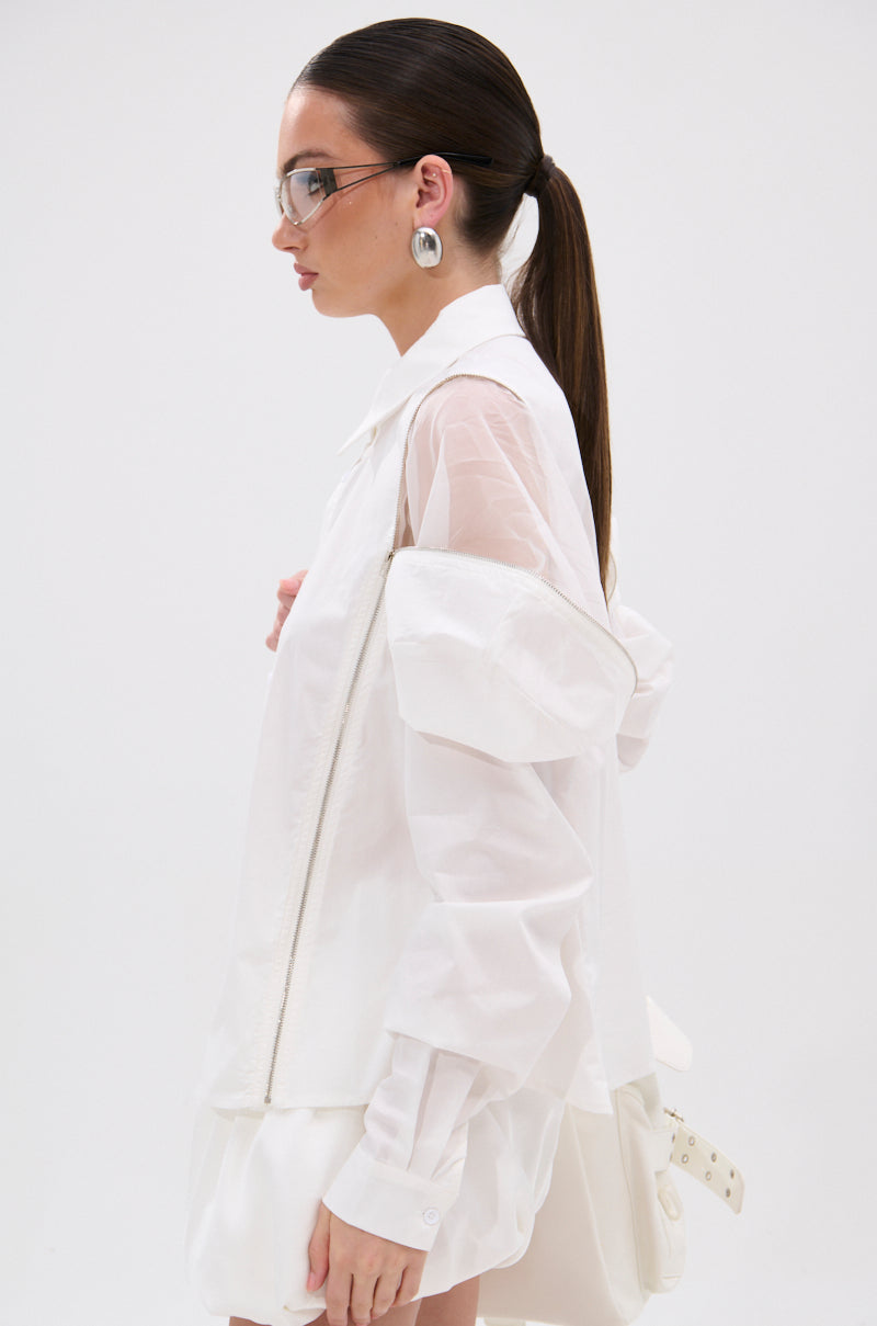 A person with long brown hair in a low ponytail wears translucent glasses, large round earrings, and the UNZIP ME BLOUSE IN WHITE—an edgy, oversized blouse with puffy sleeves and sheer fabric—standing in profile against a plain white background. 3