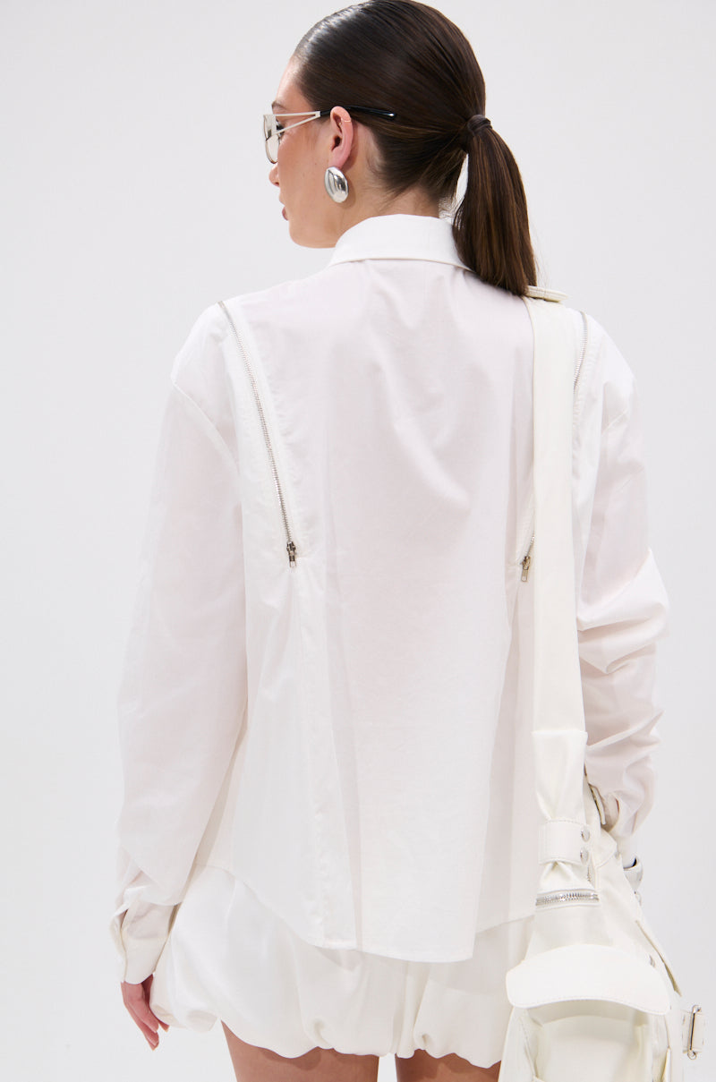 A woman with dark hair in a low ponytail, wearing large silver earrings and the UNZIP ME BLOUSE IN WHITE with functional zippers, stands facing away in a white skirt, holding a white bag over her shoulder against a plain background. 4