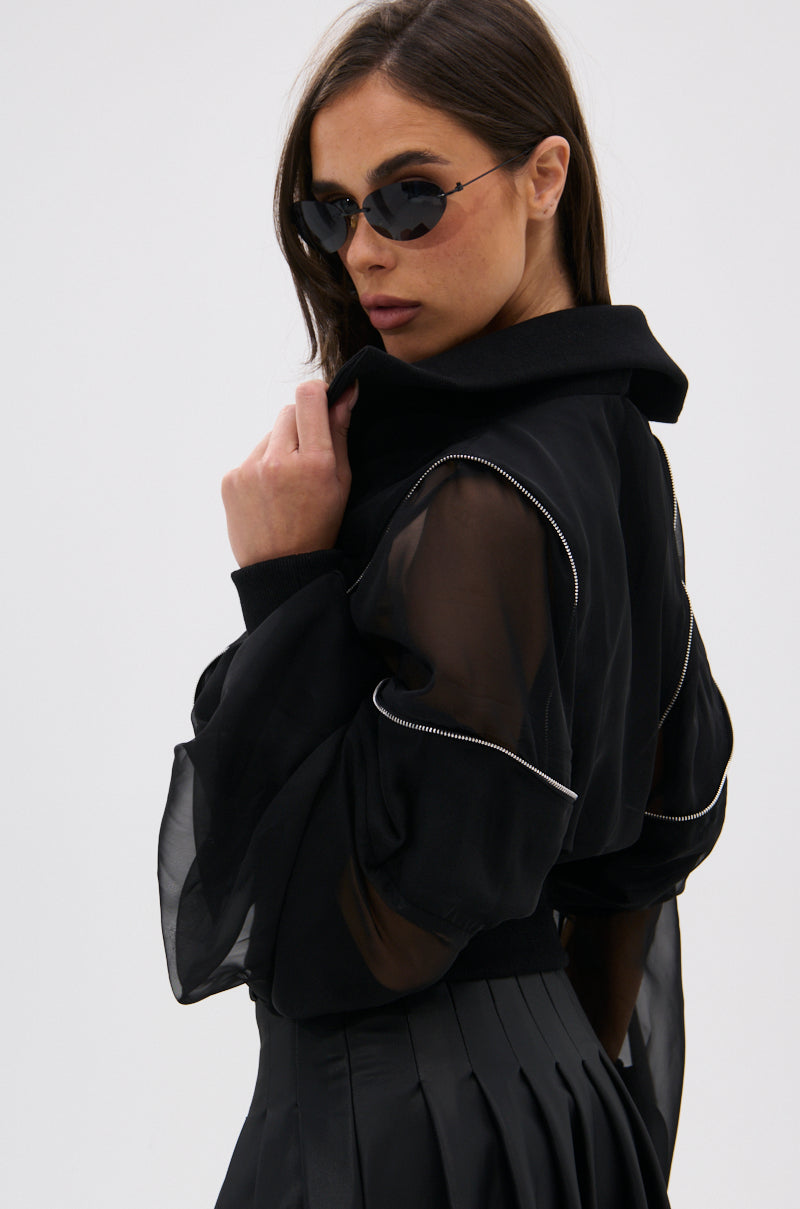 A woman in dark sunglasses and the CHOOSE YOUR FIGHTER MESH INSERT BOMBER with white piping glances over her shoulder. She styles it with a black pleated skirt, creating an elevated street look against a plain light background. 10