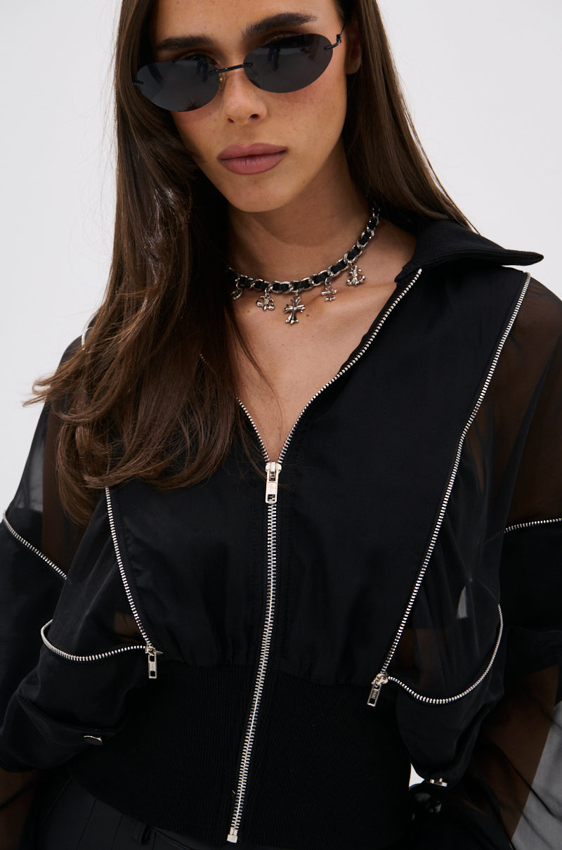 A woman stands against a plain light background, styled in black sunglasses and the CHOOSE YOUR FIGHTER MESH INSERT BOMBER with metallic zippers, plus a chunky silver flower necklace for an elevated street-style vibe. 11