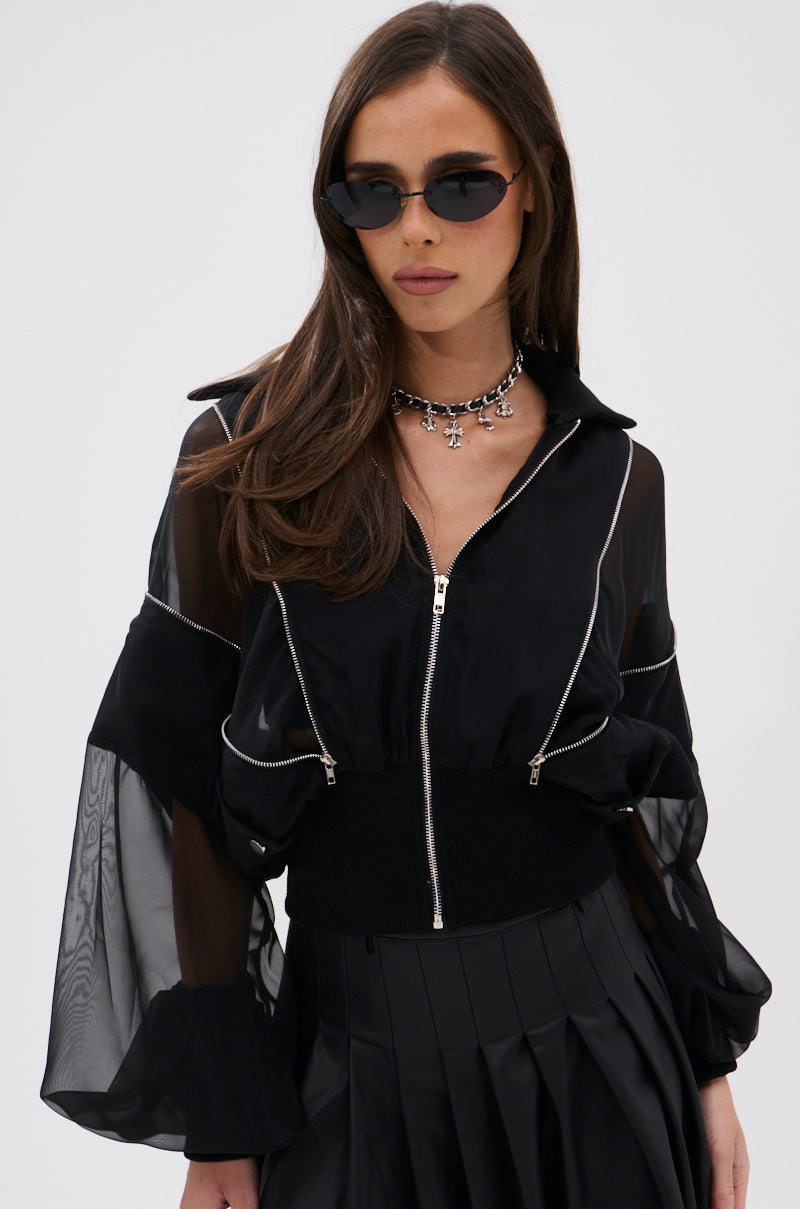 A woman with long brown hair wears black sunglasses, the CHOOSE YOUR FIGHTER MESH INSERT BOMBER with flared sleeves, a pleated black skirt, and a silver charm choker, channeling elevated street-style against a plain light background. 1