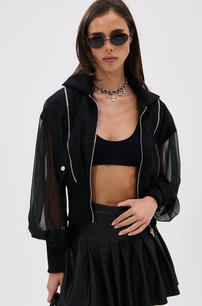 A woman in black sunglasses, a CHOOSE YOUR FIGHTER MESH INSERT BOMBER with sheer sleeves, a black bralette, pleated black mini skirt, and silver necklace stands against a plain white background. 5