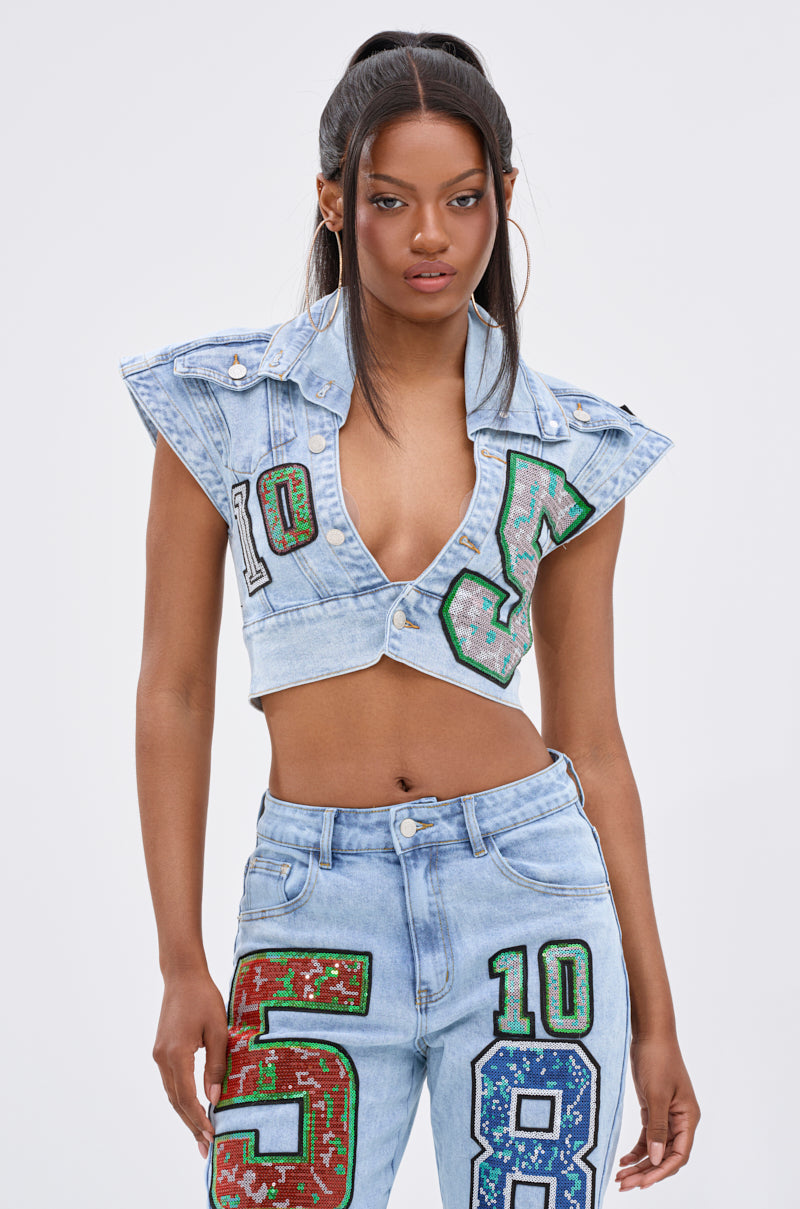 A woman wears the CHAMPION GRAPHIC DENIM BLOUSE, a sleeveless, bold-shouldered vest with a deep V-neck and colorful number patches, paired with matching high-waisted jeans for a varsity-inspired look.