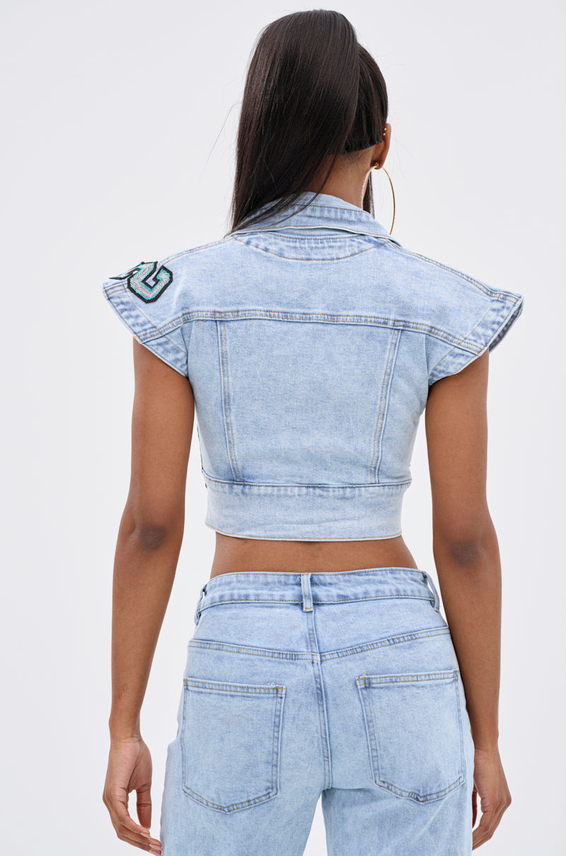 A woman is seen from the back wearing the CHAMPION GRAPHIC DENIM BLOUSE—a sleeveless, cropped, light blue denim top with Varsity Spirit patch details—paired with matching jeans against a plain light background.