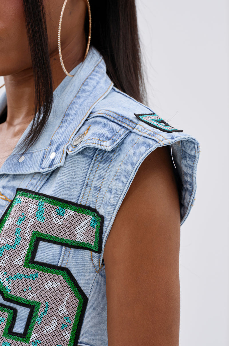 A close-up shows a person wearing the CHAMPION GRAPHIC DENIM BLOUSE with bold green and silver varsity-inspired sequins, styled with a gold hoop earring. Their face is partially out of frame.