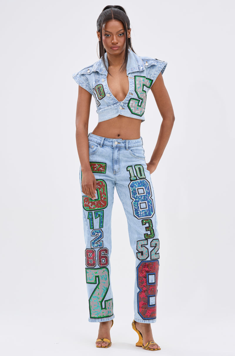 A woman showcases street-style energy in CHAMPION GRAPHIC DENIM PANT with bold, colorful numbers and sequin graphics, paired with a cropped vest and yellow heeled sandals, all set against a plain white background.