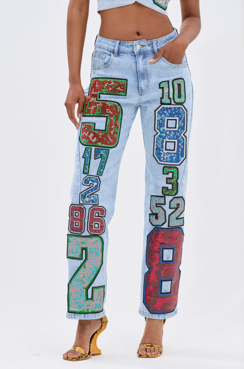 A person showcases bold street style in CHAMPION GRAPHIC DENIM PANT—light blue jeans with colorful numbers and sequin graphics—paired with yellow heeled sandals and a gray crop top, set against a white background.