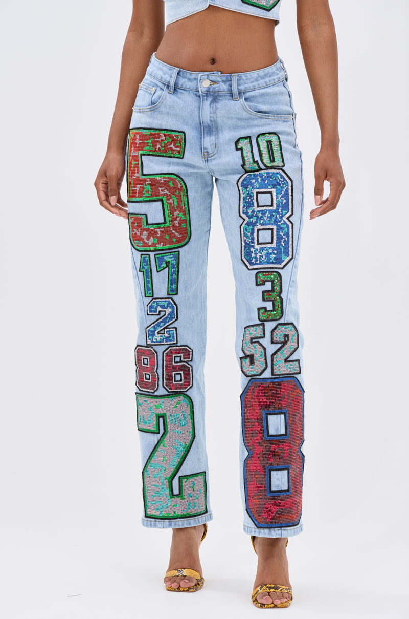 Wearing the CHAMPION GRAPHIC DENIM PANT with bold, colorful numbers, paired with yellow patterned heels and a cropped top, this street-style look pops against a plain white background.