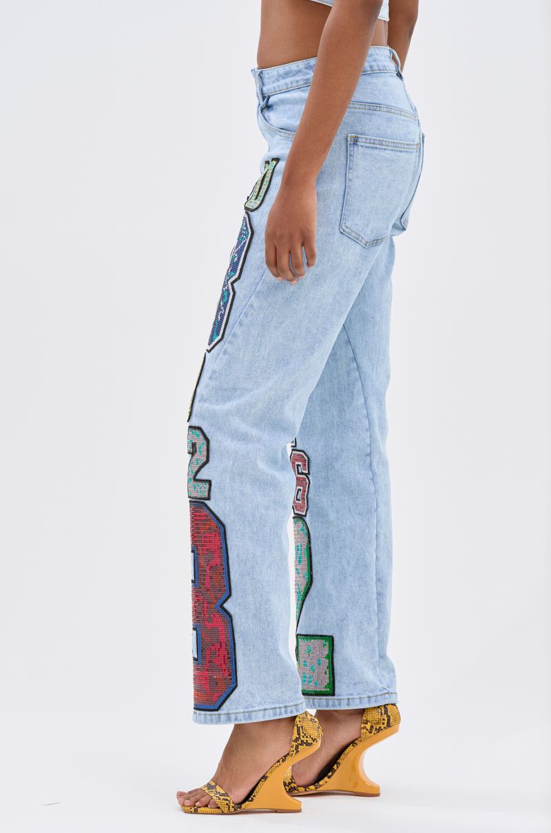 A person in CHAMPION GRAPHIC DENIM PANT with bold number patches, styled with yellow snakeskin high-heeled sandals, exudes vintage varsity vibes, shown in profile against a plain white background.