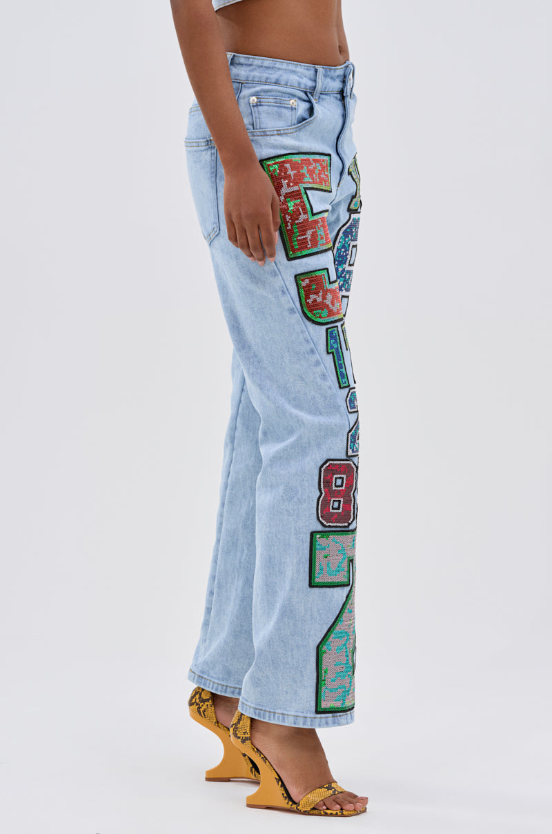 Wearing the CHAMPION GRAPHIC DENIM PANT with sequin embellishments and yellow snakeskin-patterned heeled sandals, this look radiates street-style energy against a plain white background.