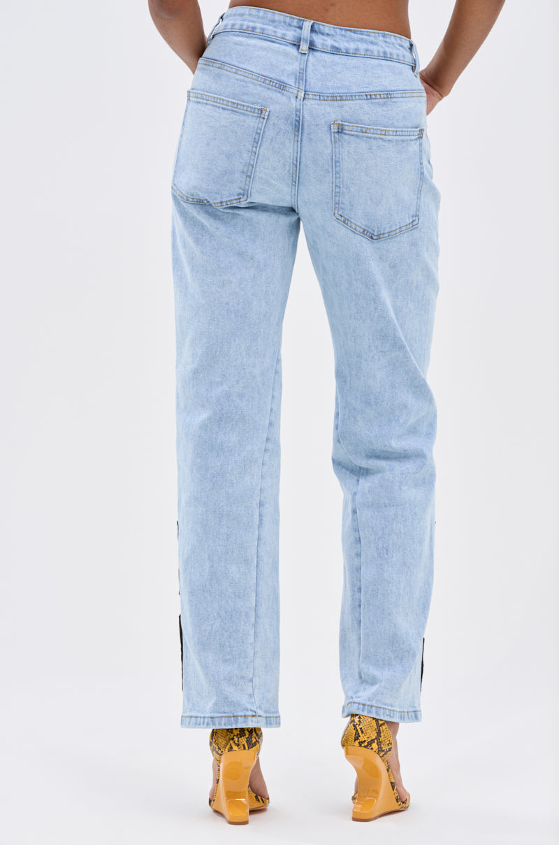 A person faces away, wearing CHAMPION GRAPHIC DENIM PANT in light blue and yellow snakeskin high-heeled sandals against a white background, exuding effortless street-style energy.