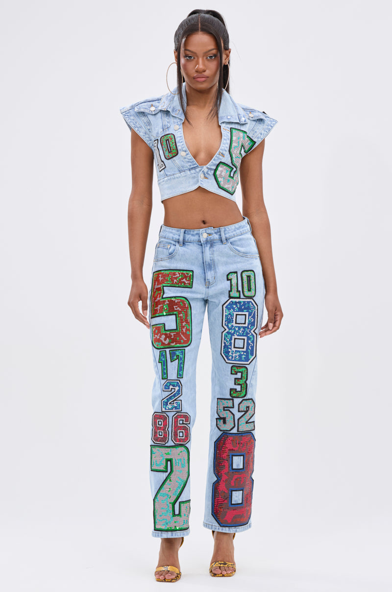 A woman models CHAMPION GRAPHIC DENIM PANT—a pair of high-waisted jeans with bold, colorful numbers—paired with a light denim crop top, capturing vintage varsity style against a plain white background.