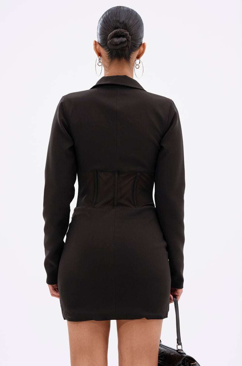 A person with dark hair in a sleek bun is shown from the back, wearing the A LITTLE BIT OF BUSINESS BLAZER DRESS—a fitted black long-sleeve blazer dress with a corset-style waist detail—and holding a small black handbag.