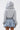 A person faces away from the camera in the FELT CUTE SWEATSHIRT DENIM MINI DRESS, featuring a gray hooded top and pleated blue denim skirt. Crisscross ribbon thigh tattoos stand out against a plain white background.