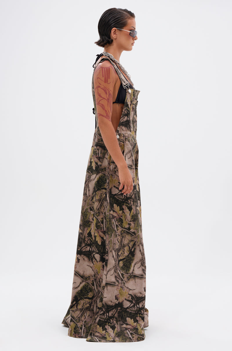 A person with slicked-back hair and sunglasses stands in profile against a plain white background, showcasing a large abstract upper-arm tattoo while wearing the CARMINE CAMO OVERSIZED OVERALLS over a black top.