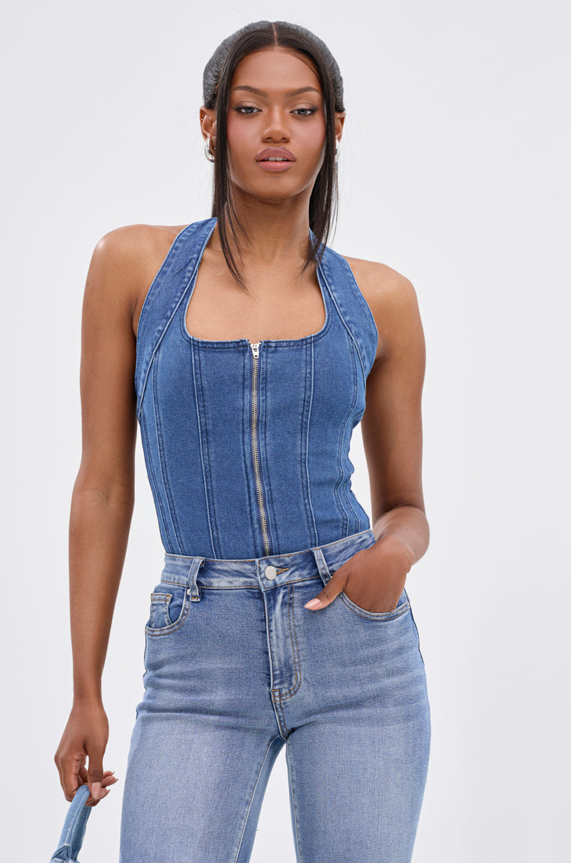 A woman stands against a plain white background wearing the CALM AND COLLECTED CORSET TOP in dark-wash denim and high-waisted light blue jeans, with one hand in her pocket.