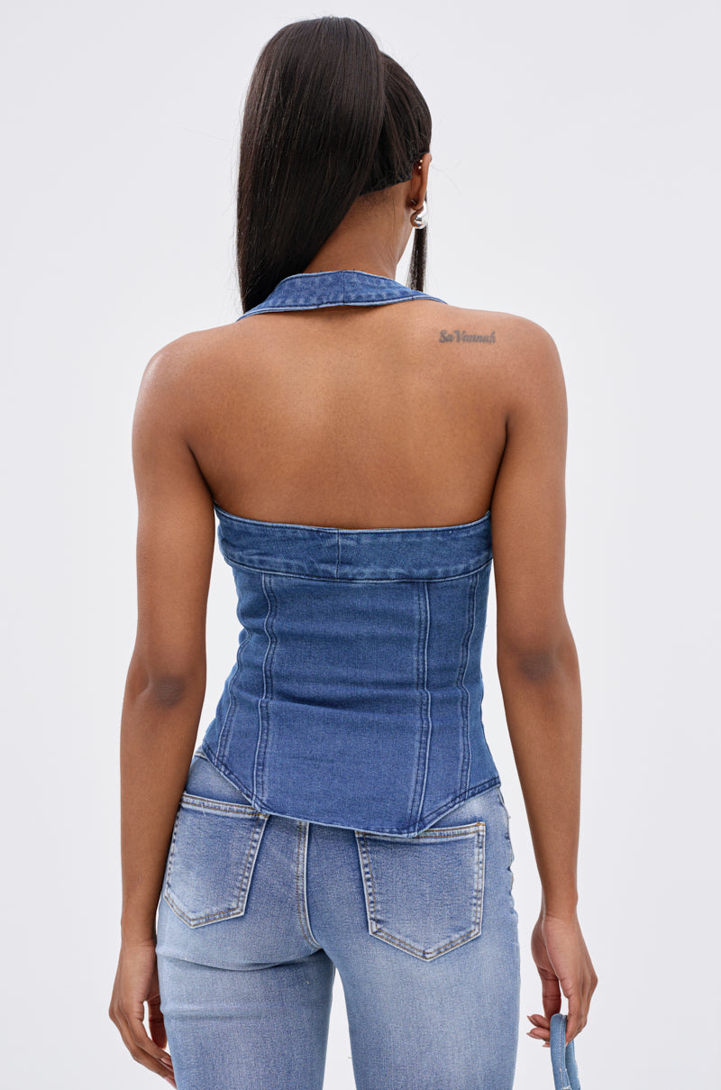 A woman models the CALM AND COLLECTED CORSET TOP, a sleeveless, strapless dark-wash denim piece, with blue jeans. Seen from behind, her long straight hair reveals a tattoo on her upper right shoulder blade. The background is plain and light-colored.