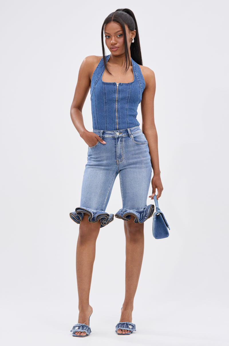 A woman stands confidently in the CALM AND COLLECTED CORSET TOP, light blue knee-length denim shorts with ruffled hems, matching heeled sandals, and a small blue handbag. Her straight hair is styled in a high ponytail.