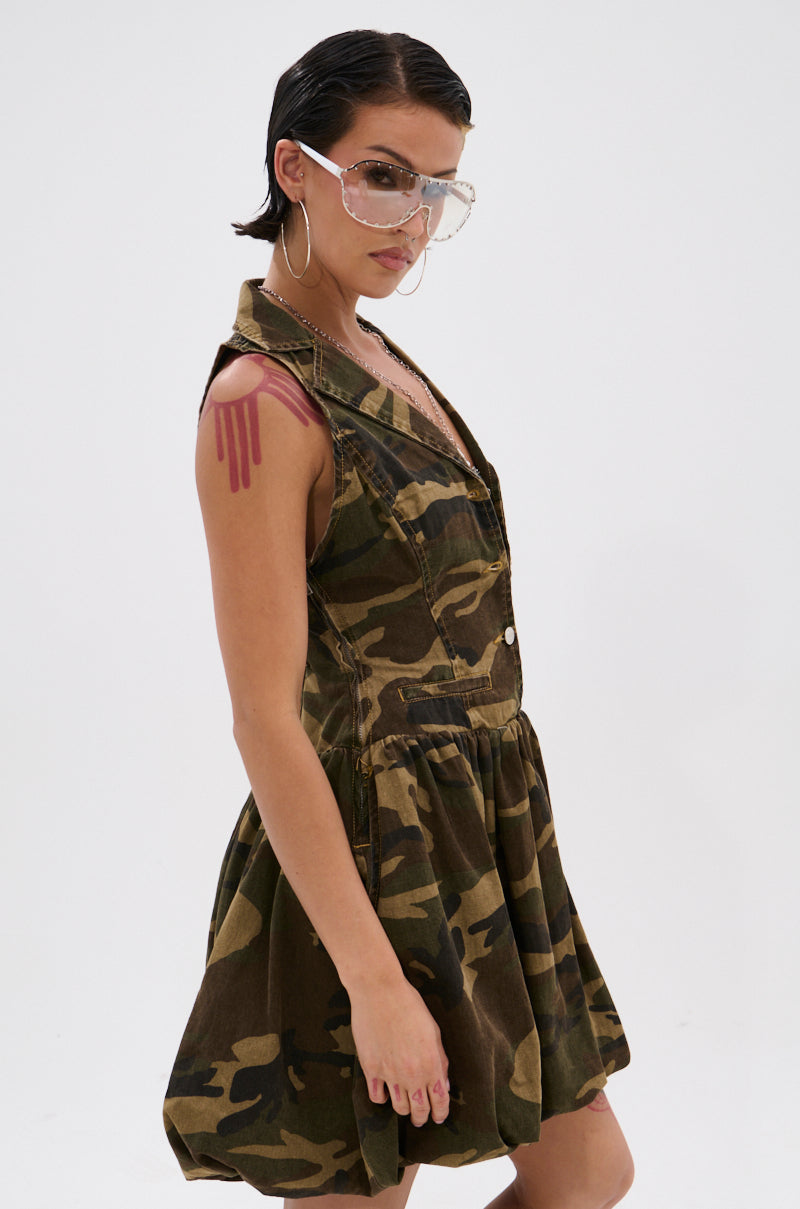 A woman models the KELLY CAMO DENIM MINI DRESS with large clear hoop earrings and transparent visor glasses, her slicked-back dark hair and pink shoulder design standing out against a plain white background.