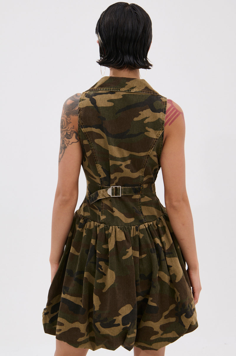 A person with short dark hair and visible arm tattoos is seen from the back wearing the sleeveless, belted KELLY CAMO DENIM MINI DRESS against a white background.