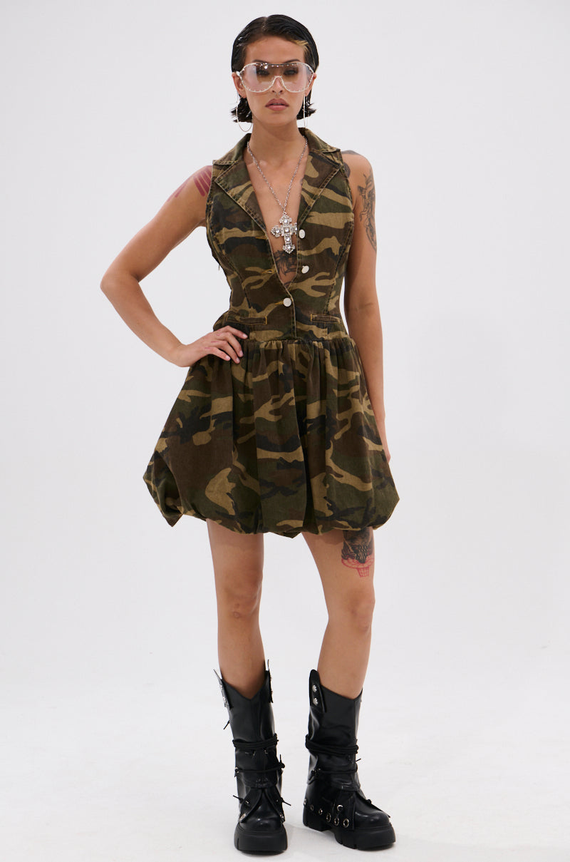 A person confidently poses in the KELLY CAMO DENIM MINI DRESS, paired with layered necklaces, sunglasses, and black combat boots, against a plain white background.