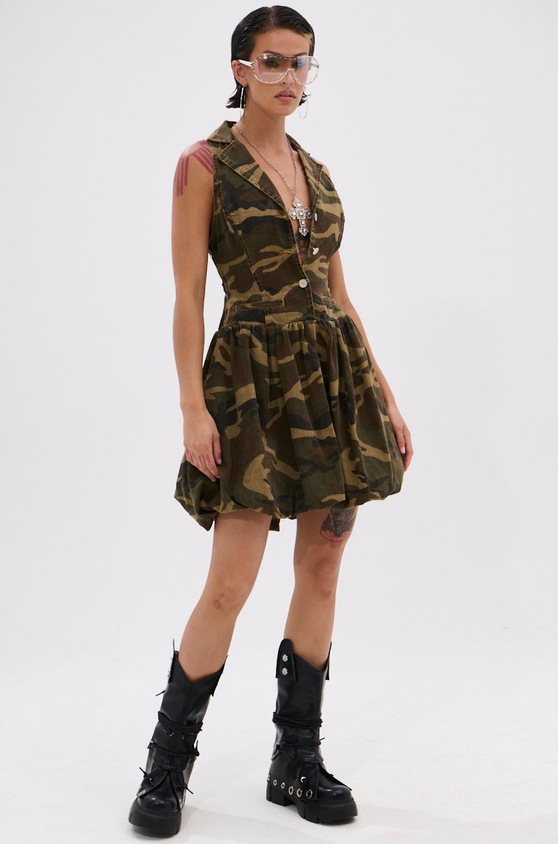 Wearing the KELLY CAMO DENIM MINI DRESS, tall black boots with metal accents, layered necklaces, and large tinted glasses, a person stands confidently against a plain white background.