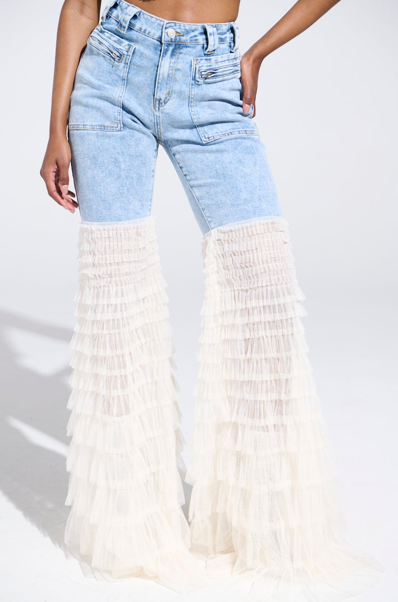 Someone is wearing the LOOK AT ME GO DENIM PANT WITH RUFFLE BOTTOM, showcasing a standard top and ruffled, sheer white fabric from mid-thigh to floor for a striking flared leg. The plain white background highlights their creative design.