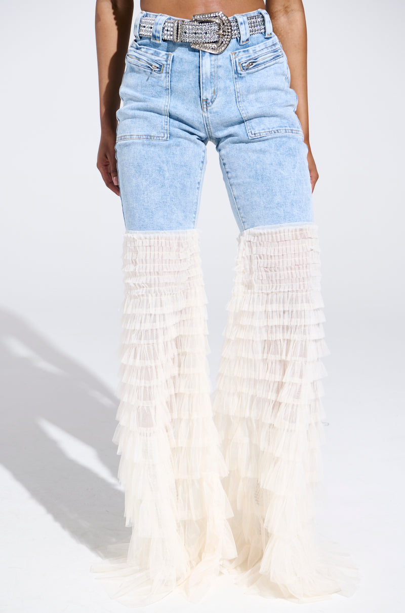 Wearing the "LOOK AT ME GO DENIM PANT WITH RUFFLE BOTTOM," one showcases high-waisted, light blue denim pants adorned with cascading cream-colored ruffled tulle from mid-thigh to ankle, featuring a flared silhouette enhanced by a decorative belt and pockets against a simple white backdrop.
