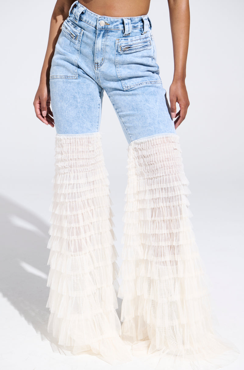 A person models the "LOOK AT ME GO DENIM PANT WITH RUFFLE BOTTOM," featuring high-waisted blue denim and flared legs with sheer layered cream tulle from knee to floor, against a plain white studio backdrop.