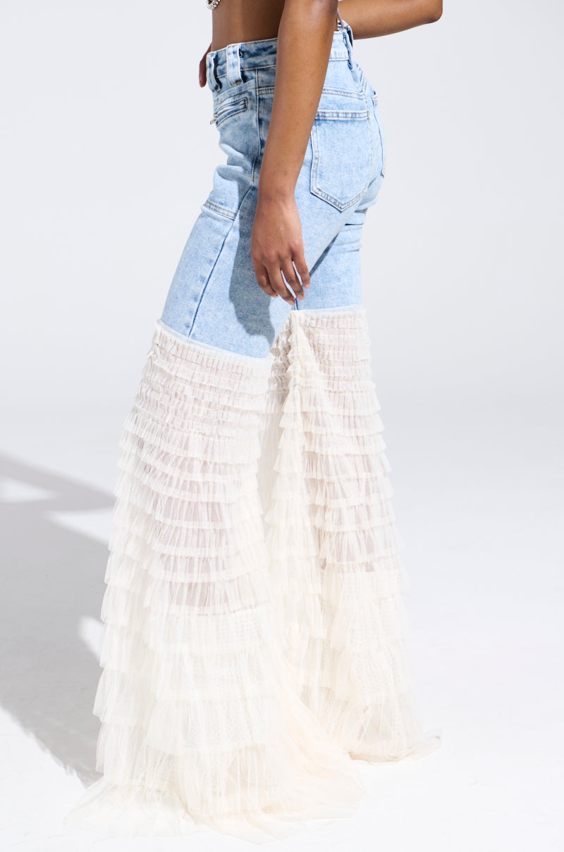 Wearing the "LOOK AT ME GO DENIM PANT WITH RUFFLE BOTTOM," a person dons light blue flared denim with layered white tulle from the knee down, posing against a white background. The focus is on the lower half of the body.