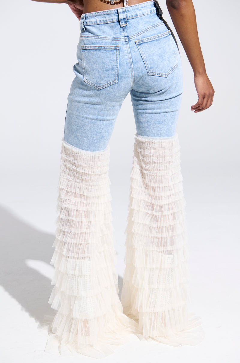 Someone is wearing the LOOK AT ME GO DENIM PANT WITH RUFFLE BOTTOM. The light blue jeans feature tulle flares from the knee down, and they are standing on a white background, facing away from the camera.