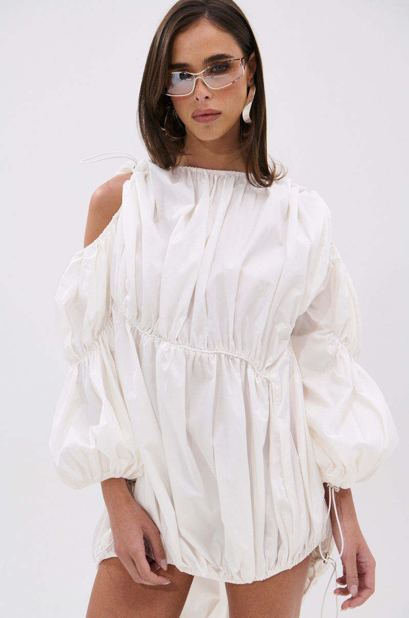A woman in the INSTA SAW IT FIRST ADJUSTABLE MINI DRESS with voluminous sleeves, paired with clear angular sunglasses and large earrings, stands against a plain white background.