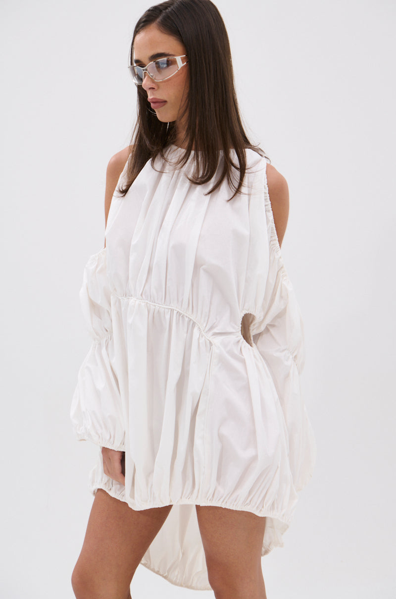 A woman with straight brown hair wears clear glasses and the INSTA SAW IT FIRST ADJUSTABLE MINI DRESS, a loose white cotton-blend design with gathered hems. She stands against a plain white background, looking to the side with her arms behind her back.