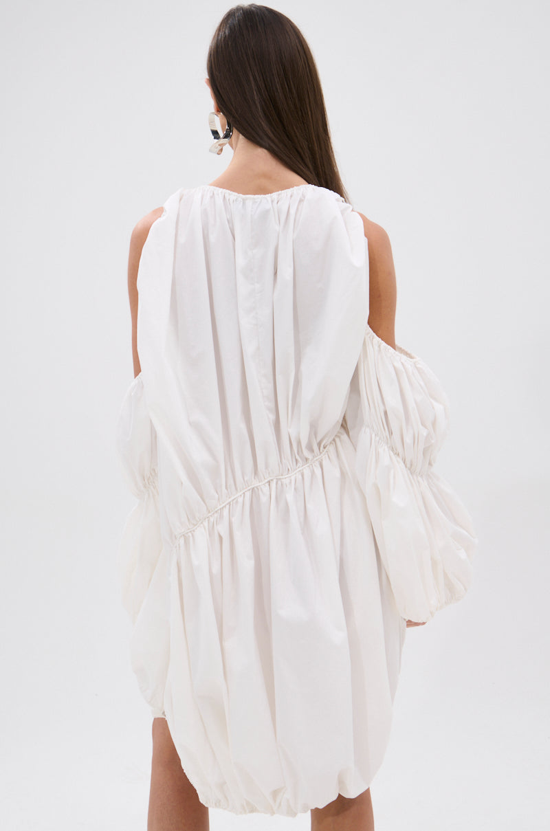 A woman with long brown hair, seen from behind, wears the INSTA SAW IT FIRST ADJUSTABLE MINI DRESS—a loose white mini dress with gathered fabric and large puffed sleeves. The background is plain white.