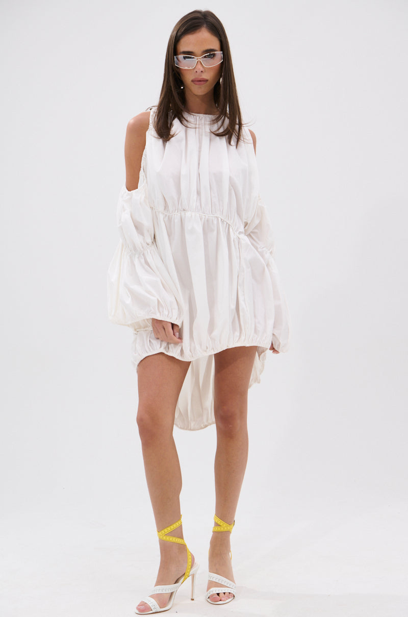 Woman wearing the INSTA SAW IT FIRST ADJUSTABLE MINI DRESS—an off-shoulder, loose white mini dress with balloon sleeves—styled with white high-heeled sandals, yellow ankle straps, and translucent glasses for a chic summer look.