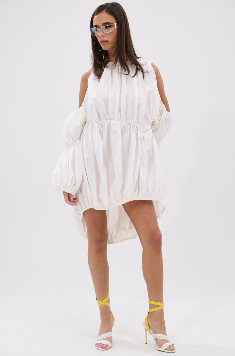 She confidently poses in the INSTA SAW IT FIRST ADJUSTABLE MINI DRESS—white, loose, with cold shoulder cutouts—styled with yellow-strapped white heels and transparent glasses for a selfie-ready look against a plain white background.